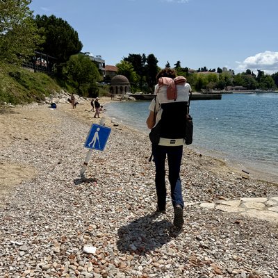 Martina von hinten auf einem Kiesstrand an der dalmatinischen Küste. Sie trägt einen grossen weiss-rosa Wanderrucksack. Links neben ihr steht ein blaues Schild mit weissem Gehenden-Piktogramm – ein speziell für sehbehinderte Personen ausgewiesener Strandabschnitt. Das Meer liegt ruhig und türkisblau im Hintergrund. Bäume, ein rundes Pavillongebäude und vereinzelte Badegäste sind in der Ferne zu sehen. Der Himmel ist strahlend blau.