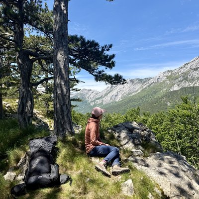 Martina sitzt seitlich auf einem Felsen und schaut in die Ferne. Sie trägt eine rosafarbene Jacke, eine beige Mütze und blaue Wanderhosen. Ihr schwarzer Rucksack liegt neben ihr auf dem Boden. Laubbäume rahmen die Szene ein. Im Hintergrund öffnet sich ein weites Tal mit den kargen, felsigen Gipfeln des südlichen Velebit-Gebirges unter blauem Himmel.