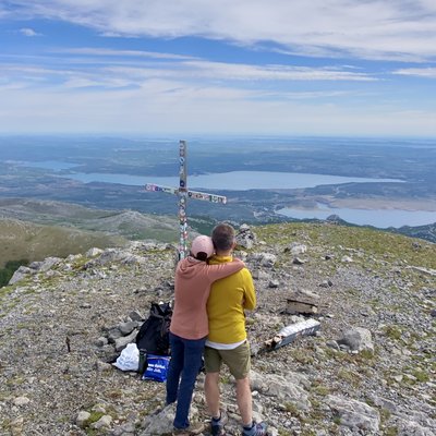 Martin und Martina stehen von hinten am Gipfelkreuz des Sveto Brdo (1710 m) und umarmen sich. Martina trägt eine rosafarbene Jacke und eine beige Mütze, Martin ein leuchtend gelbes T-Shirt und kurze Hosen. Das metallene Gipfelkreuz ist mit Plaketten und kleinen Gegenständen verziert. Der Gipfel ist karg und steinig. Im Hintergrund erstreckt sich die dalmatinische Küste mit dem blauen Adriatischen Meer bei Zadar, weit unter ihnen. Der Himmel ist hellblau mit weissen Quellwolken.