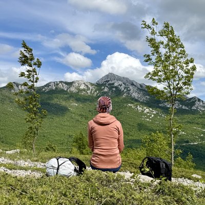 Martina sitzt von hinten auf einer offenen Karstwiese mit niedrigem Bewuchs. Sie trägt eine rosafarbene Jacke und eine bunte Mütze. Links und rechts neben ihr liegen die Wanderrucksäcke auf dem Boden. Vor ihr stehen zwei junge Einzelbäume. Im Hintergrund erhebt sich der breite, felsige Rücken des Crnopac-Gipfels mit grünen Hängen und hellgrauen Kalksteinfelsen. Der Himmel ist teils bewölkt.