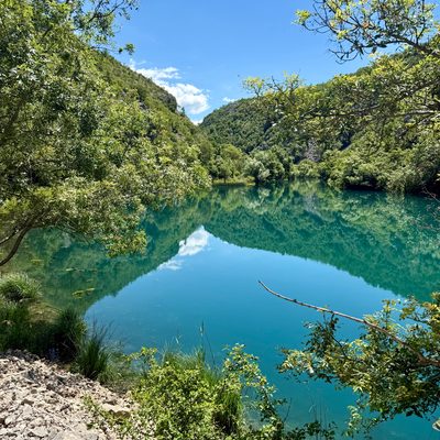 Blick durch dichtes grünes Laub auf einen ruhigen, türkis-grünen Flussabschnitt der Krupa-Schlucht. Das Wasser ist so still, dass es die bewaldeten Berghänge und den hellblauen Himmel mit wenigen weissen Wolken klar spiegelt. Am linken Bildrand schiebt sich eine kleine Geröllhalde ins Bild, im Vordergrund Zweige und Sträucher. Hinter dem Wasser steigen die steilen, dicht bewachsenen Hänge der Schlucht steil auf.