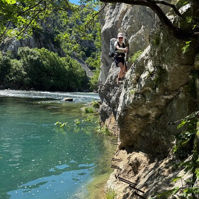 Martina steht auf einer schmalen Felsstufe an einer steilen, hellen Kalksteinwand und blickt mit einem Lächeln in die Kamera. Sie trägt einen grossen blau-grauen Wanderrucksack, ein weisses Top und eine Cap. Unter ihren Füssen führen schmale Holzbohlen und Stahlseile als Sicherung an der senkrechten Wand entlang. Links unten leuchtet das türkis-grüne Wasser der Krupa, gegenüber steigen dicht bewaldete Schluchtwände auf. Im Vordergrund rahmen Zweige und Blätter eines Baumes die Szene ein.