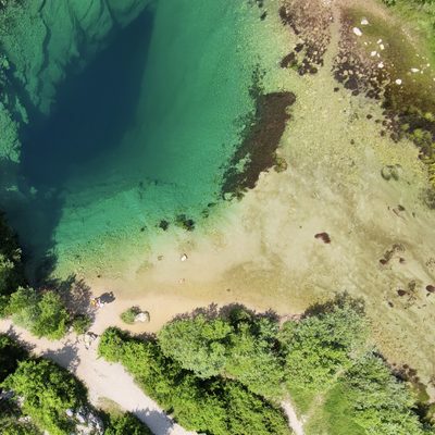 Drohnenaufnahme aus etwa 30 Metern Höhe direkt über der Cetina-Quelle. Das fast kreisrunde Quellbecken zeigt im Zentrum tiefdunkles Blau, das nach aussen in Türkis und helleres Grün übergeht. Rechts und unten ziehen sich Sandbänke, Schilf und grüne Wasserpflanzen am flachen Ufer entlang. Auf dem Sandstrand unten links sind als winzige Figuren Martina und Martin zu erkennen, daneben ein Schotterweg, der durch die grüne Ufervegetation weiterführt.