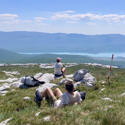 Drohnenaufnahme von einem Karstplateau auf der Dinara mit Blick weit hinunter ins Tal. Im Vordergrund liegt eine Person im Gras zwischen Karststeinen, in beigen Wanderhosen und braunen Wanderstiefeln. Etwas dahinter sitzt eine zweite Person mit Käppi auf einem Felsblock und blickt in die Ferne, ein Wanderrucksack liegt daneben. Rechts ragt eine rot-weiss gestreifte Wanderwegmarkierung in den Himmel. Weiter unten erstreckt sich der türkis-blaue Peručko See als langgezogener Streifen vor einer Kette weiter, blauer Berghöhen. Strahlend blauer Himmel mit feinen Wolken.