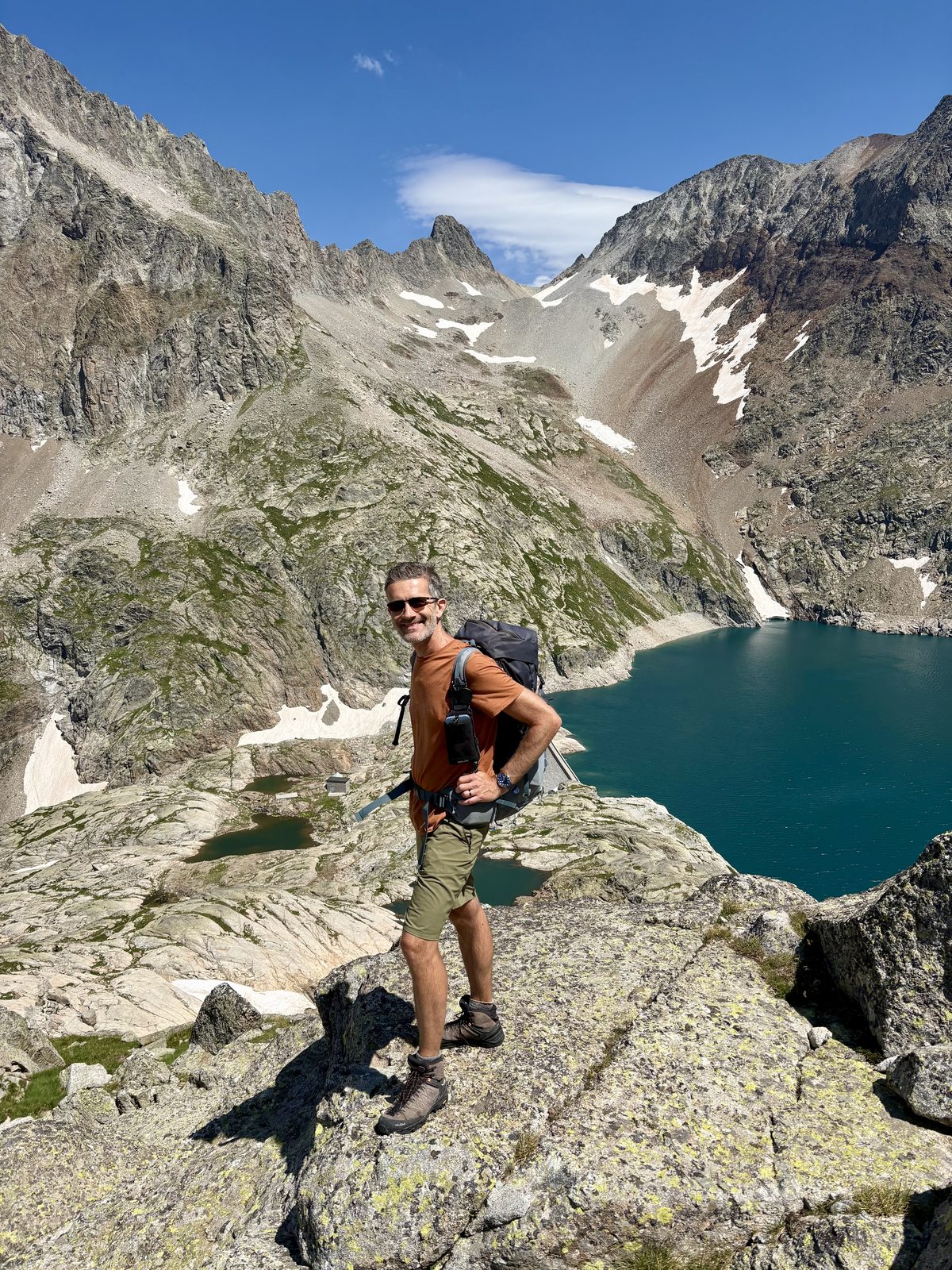 Martin steht lächelnd auf einem Felsen vor einem türkisfarbenen Bergsee. Er trägt Sonnenbrille und Rucksack. Im Hintergrund felsige Gipfel mit Schneefeldern und strahlend blauer Himmel.