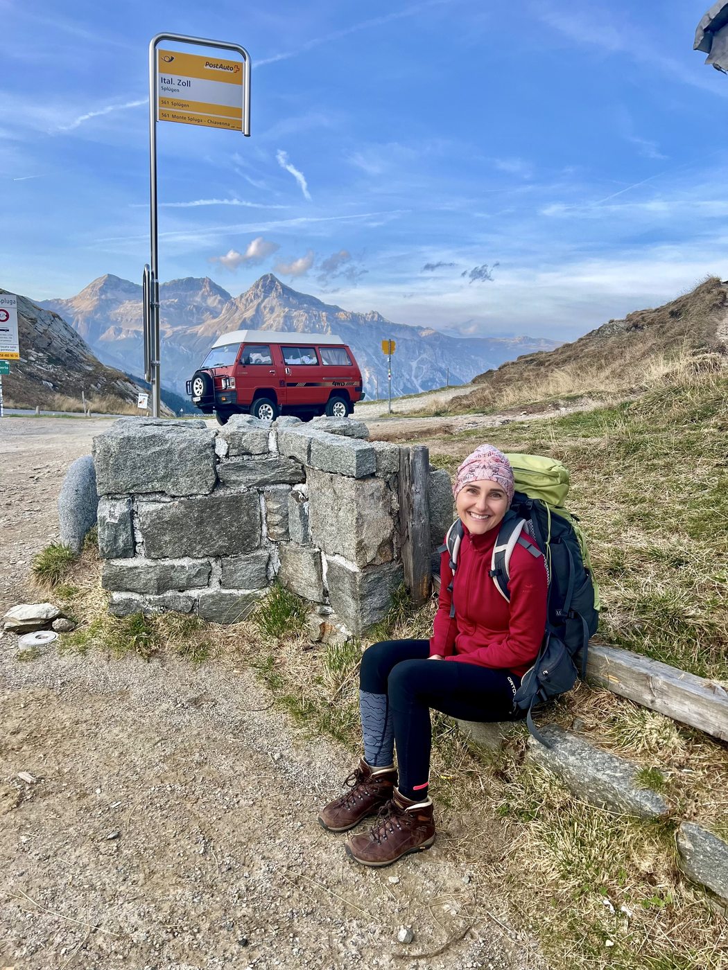 Martina sitzt mit grossem Rucksack auf einer Steinmauer an einer Postautohaltestelle auf einem Bergpass. Sie lächelt, trägt eine rosa Mütze und rote Jacke. Dahinter ein roter VW-Bus und schneebedeckte Gipfel.