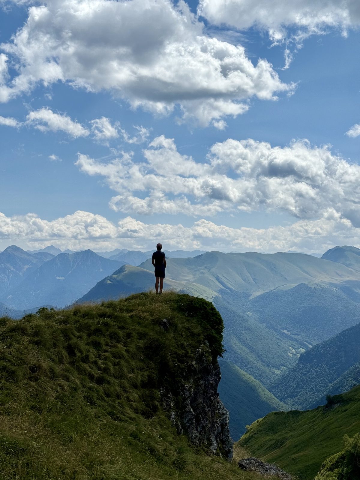 Eine Person steht als kleine Silhouette auf der Kante eines grasbewachsenen Felsvorsprungs. Unter ihr fällt das Gelände steil ab in ein bewaldetes Bergtal. Grosse Wolken türmen sich am Himmel.