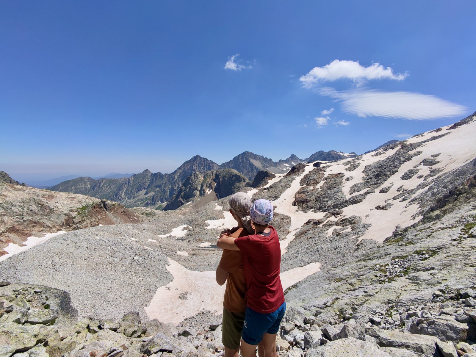 Martina und Martin umarmen sich auf einem steinigen Hochplateau und schauen ins Tal. Hinter ihnen erstreckt sich ein weites Gletscherkar mit Schneefeldern und zerklüfteten Felsgipfeln unter blauem Himmel.