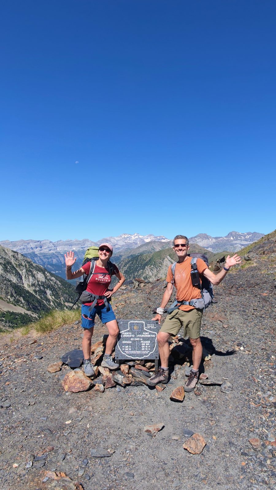 Martina und Martin winken lachend in die Kamera, beide mit Rucksäcken. Zwischen ihnen ein Grenzstein mit einer Infotafel auf einem Bergsattel. Im Hintergrund ein weites Bergpanorama unter tiefblauem Himmel.