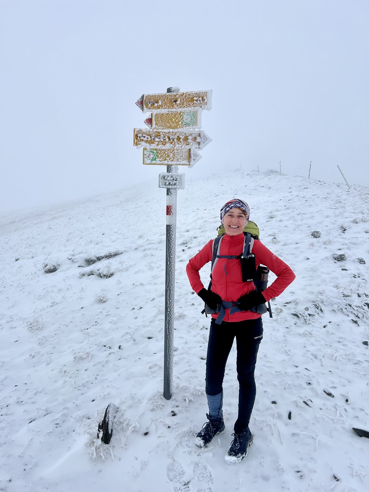 Martina steht lächelnd neben einem vereisten und schneebedeckten Wegweiser auf einem kahlen Bergkamm. Sie trägt rote Jacke, Stirnband und Rucksack. Alles ist in Nebel und Neuschnee getaucht.
