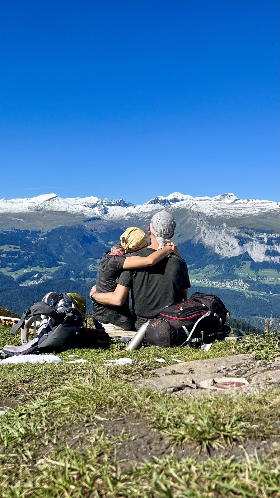 Martina und Martin sitzen Arm in Arm auf einer Almwiese, Rücken zur Kamera, und blicken auf schneebedeckte Berggipfel unter strahlendem Blau. Ihre Rucksäcke liegen neben ihnen im Gras.