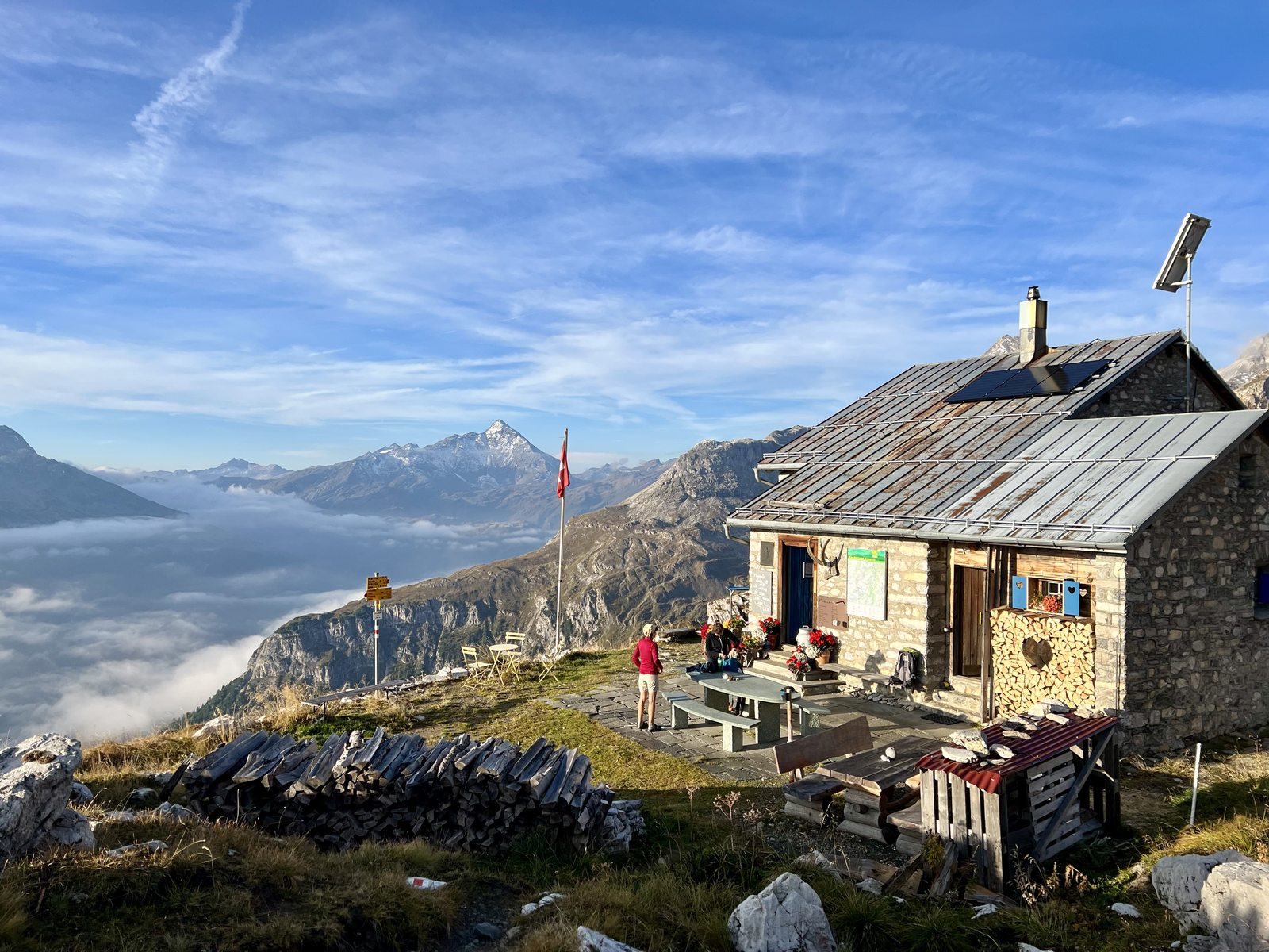 Eine rustikale Berghütte aus Stein mit Schweizer Fahne steht hoch über einem Wolkenmeer. Eine Person in roter Jacke steht auf der Terrasse davor. Im Hintergrund ragen einzelne Berggipfel aus dem Nebel.