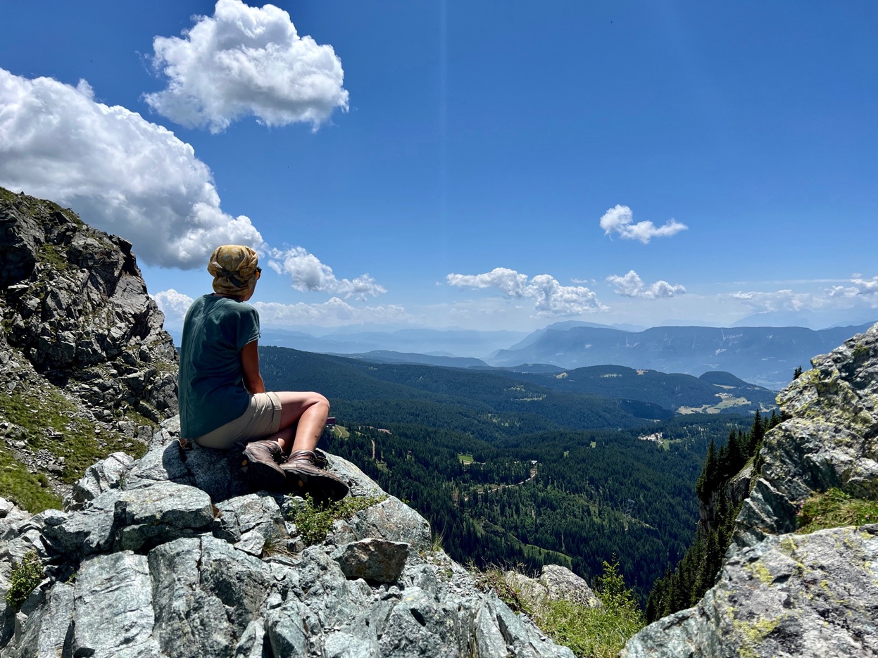 Eine Person sitzt allein auf einem Felsen, Rücken zur Kamera, und schaut in die Weite. Vor ihr erstreckt sich ein bewaldetes Tal mit einem grossen See am Horizont. Blauer Himmel mit Sommerwolken.